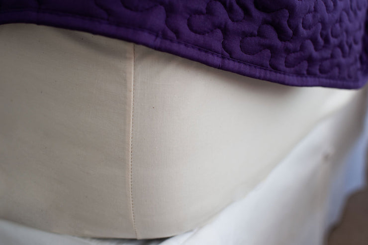 Corner of mattress made with organic cotton fitted sheet in natural color with a purple bedspread.